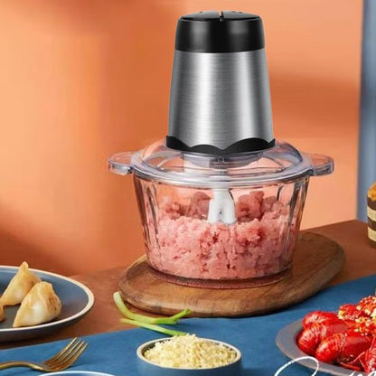 Présentation du hachoir électrique sur une table bien garnie d'aliments divers. On voit une préparation de jambon haché au travers du bol en verre.
