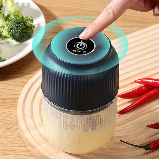 Mini hachoir USB au couvercle-moteur noir et base semi transparente présenté sur planche en bois et table en bois, entouré de brocoli et piment rouge. Vue de proche pour montrer le bouton d'activation