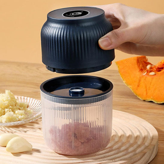 Mini hachoir USB au couvercle-moteur noir et base semi transparente présenté sur planche en bois et table en bois, entouré d'ail et courge. Vue de proche, on voit une main qui soulève le couvercle moteur et in peut voir du jambon haché à l'intérieur