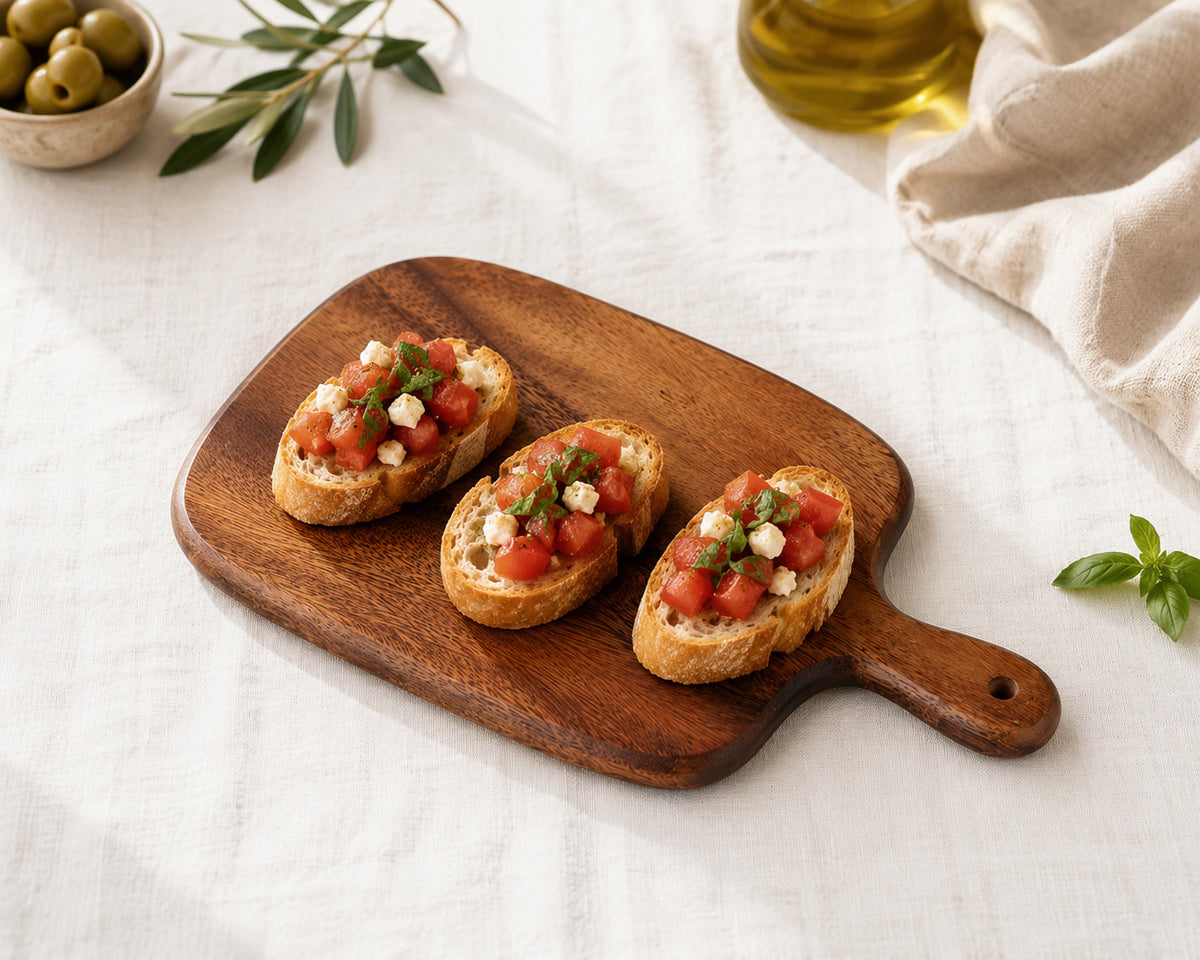 Bruschettas aux tomates et basilic présentées sur une planche à découper bois d'acacia avec huile d'olive et olives sur nappe en lin.