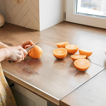 Planche à découper en acrylique clair avec son rebord plié pour s'accrocher au comptoir. L'image montre une femme coupant une orange à la lumière du soleil