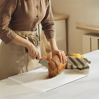 Planche à découper déposée sur un comptoir au décor champêtre montrant une femme tranchant une miche de pain avec un couteau du chef
