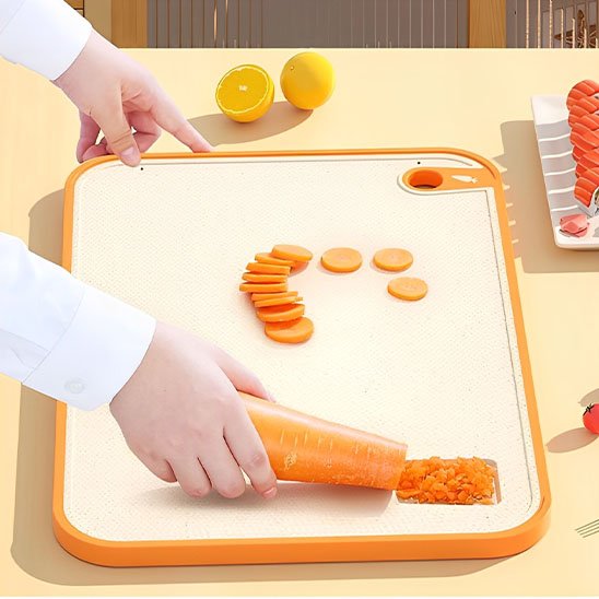 Jolie planche à découper en silicone de couleur orange  avec sa râpe incrustée, démontre une carotte râpée et tranchée en rondelles