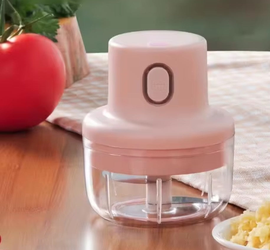 Présentation du mini hachoir 100 ml, dans un très joli décor aux tons rosés, près d'un napperon rose et blanc à carreaux, qui lui est sur une table en bois, et autour du mini hachoir, il y a de la coriandre, une tomate, et de l'ail broyé dans un petit plat blanc.
