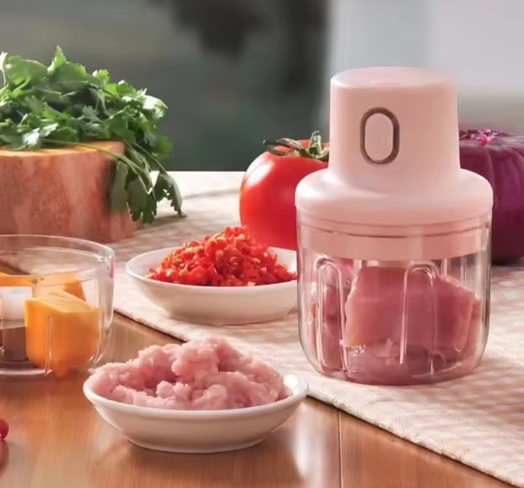 Présentation du mini hachoir 250 ml, dans un très joli décor aux tons rosés, sur napperon rose et blanc à carreaux, qui lui est sur une table en bois, et autour du mini hachoir, il y a de la coriandre, une tomate, un oignon et des aliments broyés dans des petits plats blancs